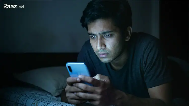 Worried young Indian man reading about premature ejaculation on his phone in bed at night